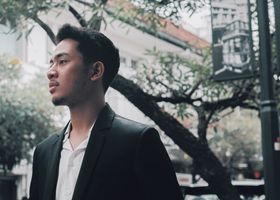 A stylish young man in a black suit stands confidently in an urban outdoor setting.